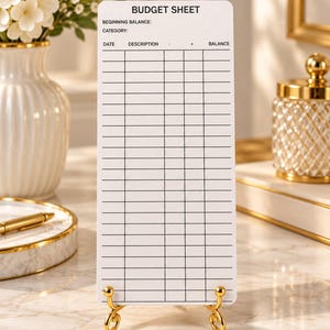 May include: A white rectangular budget sheet printed with black text and grid lines, featuring the header BUDGET SHEET, fields for beginning balance and category, and columns for date, description, minus, plus, and balance, held by a gold stand.