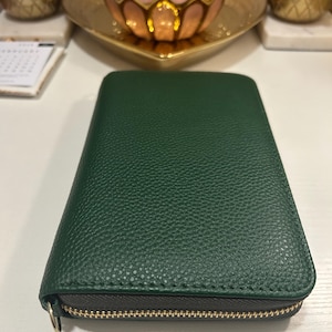 Puede incluir: Una cartera verde oscuro con una superficie texturizada y una cremallera dorada. La cartera está cerrada y parece ser de cuero. El tirador de la cremallera es un pequeño anillo dorado.