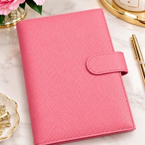 May include: A pink leather-textured planner with a matching snap-button closure strap. The rectangular cover features a pebbled finish and is positioned next to a gold pen, a white candle with the text DREAM PLAN DO, and gold paper clips.