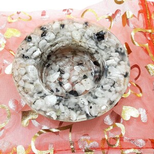 Pode incluir: Um porta-velas redondo branco e preto salpicado com um pequeno centro redondo aberto. O porta-velas está sobre uma bolsa de malha rosa e dourada com corações.