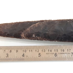 Puede incluir: Una punta de flecha de piedra de color marrón oscuro, puntiaguda, que mide aproximadamente 17,8 cm de largo. La punta de flecha está apoyada en una regla con marcas en centímetros.