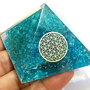 May include: A teal blue crystal pyramid with a gold flower of life symbol on the front. The pyramid is made of small, clear crystals.