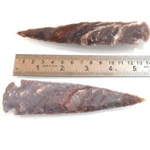 以下が含まれることがあります： 縞瑪瑙製の矢じり2本。矢じりは約13cmと15cmの長さです。矢じりは薄い茶色で、濃い茶色の縞模様が入っています。