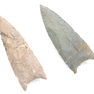 May include: Two ancient arrowheads. One is a light tan color with a rough texture, and the other is gray with a smooth, polished appearance. Both have a triangular shape with a pointed tip and a notched base.
