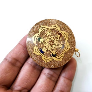 May include: Gold-toned pendant with a clear resin base and a gold-colored geometric design. The design features a flower-like shape with seven points, each containing a different colored gemstone. The pendant has a gold-colored loop for attaching a chain.