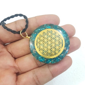 May include: A round pendant with a gold-colored Sacred Geometry design set in teal resin. The pendant hangs from a black cord. The gold design is a repeating pattern of interlocking circles. The pendant is held in a hand.