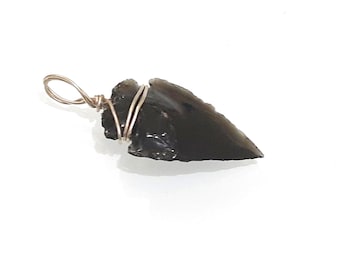 Può includere: Un ciondolo a forma di punta di freccia in ossidiana nera avvolto da un filo d'argento. La punta di freccia è di un nero opaco scuro, con una punta affilata e una base leggermente curva.
