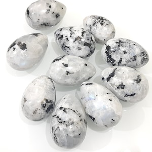 Puede incluir: Una colección de diez cristales de piedra lunar pulidos en forma de huevo. Las piedras son principalmente blancas con destellos iridiscentes e inclusiones negras. Los huevos varían en tamaño y están dispuestos sobre una superficie blanca, mostrando su belleza natural y patrones únicos.