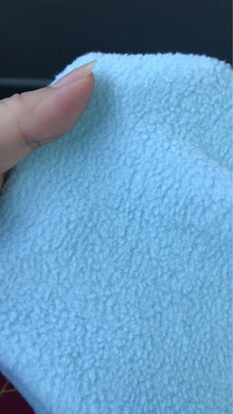 May include: Close-up of a light blue fleece fabric. The fabric has a soft, textured surface. The image shows a portion of the fabric, with a finger touching the edge. The fleece appears to be a solid color.
