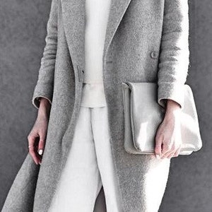 May include: A grey wool coat with a white sweater and white trousers. The coat has a long, flowing style and a simple design. The person is holding a grey clutch bag.
