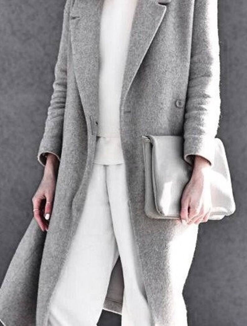 May include: A grey wool coat with a white sweater and white trousers. The coat has a long, flowing style and a simple design. The person is holding a grey clutch bag.