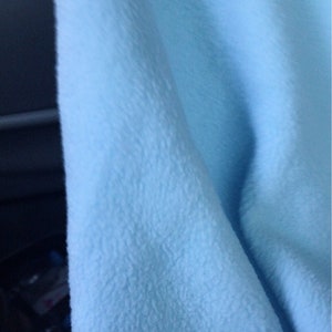 May include: Close-up of a soft, light blue fabric with a textured, plush surface. The fabric is folded, creating gentle curves and shadows. The material appears to be a cozy blanket or throw.
