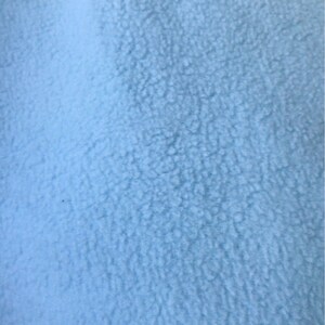 May include: Close-up of a light blue fleece fabric. The material has a soft, textured appearance with a slightly uneven surface. The color is a solid, pale blue, and the fabric appears to be thick and warm.