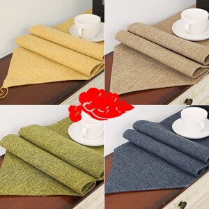 May include: Four table runners in different colours: yellow, brown, green, and blue. Each runner is made of a textured fabric and is folded in half. A white cup and saucer are placed on each runner.