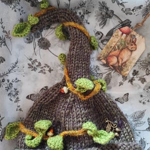 Peut inclure: Un chapeau tricoté à la main, fantaisiste, dans les tons de marron et de violet, orné de feuilles vertes et d'une vigne dorée. Le chapeau est orné d'une étiquette avec une illustration de lapin. Un article unique et artisanal.