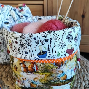Op de afbeelding: Een stoffen mand met een boslandschap print, gevuld met roze en rode bollen wol en breinaalden. De mand heeft een witte bloemenvoering en een oranje stippelrand.