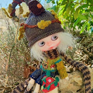Peut inclure: Une poupée artisanale sur le thème de la forêt. La poupée porte un chapeau, une écharpe et un pull tricotés dans des tons terre, associés à un sac rouge en forme de champignon et des bottes marron. La poupée est assise sur un rocher.