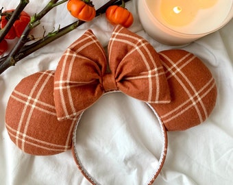 May include: A pair of brown and white plaid Minnie Mouse ears with a large bow. The ears are on a white background with small orange pumpkins.