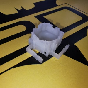 Puede incluir: Un modelo de impresión 3D en plástico blanco de un pequeño edificio con una base cuadrada y un techo cuadrado. El modelo tiene una pequeña abertura en un lado.
