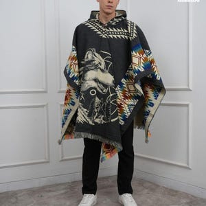 Op de afbeelding: Een donkergrijze poncho met capuchon en een wolfontwerp. De poncho heeft een geometrisch patroon in blauw, rood en crème langs de randen. De wolfafbeelding is in crème en zwart. De poncho heeft een gefranjerde zoom.