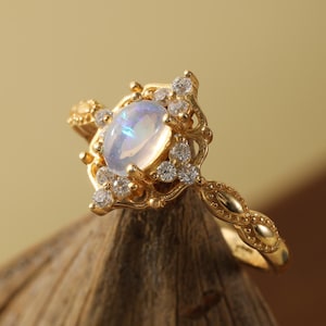 May include: A gold ring featuring a central oval-shaped, iridescent gemstone, surrounded by smaller, clear gemstones. The ring has an ornate, filigree design with a twisted band. The ring is displayed on a piece of wood.