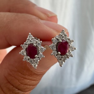 Op de afbeelding: Een paar oorstekers met ovale, dieprode edelstenen, omringd door kleinere, heldere edelstenen. De oorbellen hebben een sterrenpatroon en zijn gezet in een zilverkleurig metaal.