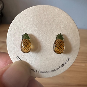May include: A pair of pineapple stud earrings. Each earring features a detailed, yellow and brown pineapple design with a green top. The earrings are displayed on a round, off-white card with text that reads "usa / handmade in California".