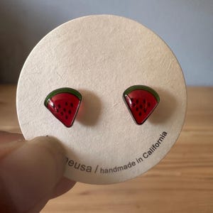 May include: A pair of watermelon slice stud earrings. Each earring features a red, triangular design with black seed details and a green rind. The earrings are set in a silver-colored metal and are displayed on a round, off-white card.