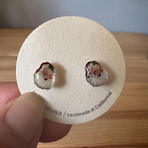 May include: A pair of Santa Claus stud earrings. Each earring features a miniature image of Santa with a red hat, white beard, and rosy cheeks. The earrings are displayed on a round, off-white card.
