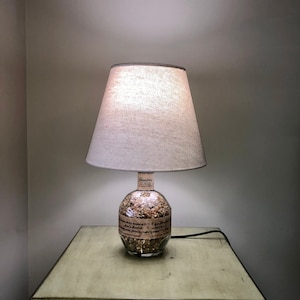 Puede incluir: Una pantalla de lámpara blanca con una pantalla de tela beige se encuentra sobre una mesa de madera marrón claro. La base de la lámpara es una botella de vidrio transparente llena de pequeñas piedras. La botella tiene una etiqueta que dice "Blenders Whiskey."