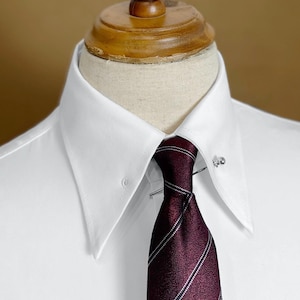 Op de afbeelding: Close-up van een wit overhemd met een gespreide kraag en een bordeauxrode stropdas met witte strepen. Een zilveren stropdaspen houdt de stropdas vast. Het overhemd wordt op een paspop getoond.