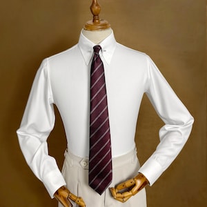 May include: A white dress shirt with a spread collar and a burgundy striped tie. The tie is secured with a tie bar. The shirt is paired with beige dress pants. The shirt has long sleeves and buttoned cuffs.
