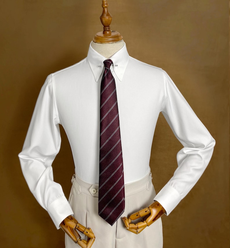 May include: A white dress shirt with a spread collar and a burgundy striped tie. The tie is secured with a tie bar. The shirt is paired with beige dress pants. The shirt has long sleeves and buttoned cuffs.