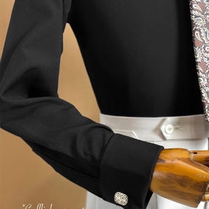 May include: A black long-sleeve shirt with a white button-up collar and cuffs. The cuffs feature silver cufflinks. A patterned tie is visible on the right side. The text "* Cufflinks are not included" is at the bottom.