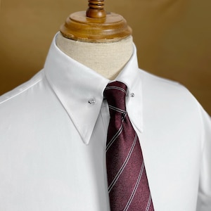 May include: A white dress shirt with a burgundy tie featuring thin white stripes. The tie is secured with a silver collar pin. The shirt is displayed on a mannequin.