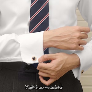 May include: A close-up of a person wearing a white dress shirt with a navy blue tie with red stripes. The person is wearing gold cufflinks with a black center. The text "Cufflinks are not included" is at the bottom.
