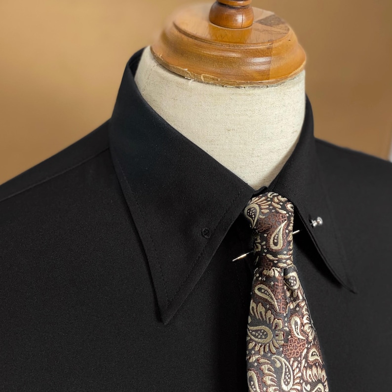May include: Close-up of a black dress shirt with a wide collar and a patterned tie. The tie features a paisley design in shades of brown, tan, and green. A silver tie pin secures the tie to the shirt.