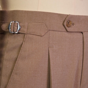 May include: Light brown trousers featuring a high waistband with a side adjuster buckle, a single button closure, and a vertical pleat. A maroon label on the waistband displays the text HIEU DO 90s Classic menswear Since 2020.