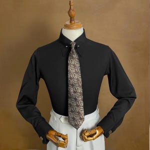 May include: A black long-sleeve shirt with a classic collar and a patterned tie. The tie features a brown and cream paisley design. The shirt is paired with white pants and is displayed on a mannequin.