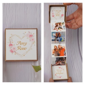 May include: A small, brown box with a white lid decorated with pink flowers and gold lettering. The lid opens to reveal a series of photos with the text "I can't say I do without you" and "Will you be my bridesmaid?"