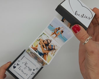 Puede incluir: Una caja negra con un corazón blanco y la palabra "besties" en la parte superior. La caja está abierta y se saca una tira de fotos. Las fotos muestran a dos mujeres sonriendo y divirtiéndose. La caja también tiene una etiqueta blanca con el texto "Always & Forever" y corazones.
