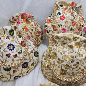 Op de afbeelding: Verschillende crèmekleurige tassen met trekkoord en ingewikkelde bloemenborduurwerken in verschillende kleuren, waaronder rood, roze en goud. Elke tas heeft een kralenhandvat en een decoratieve kwast.