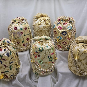 Peut inclure: Six sacs à cordon brodés, ornés de motifs floraux et géométriques. Les sacs sont principalement de couleur crème avec des accents dorés, rouges, bleus et verts. Chaque sac est orné d'une bordure de perles et de glands dorés.