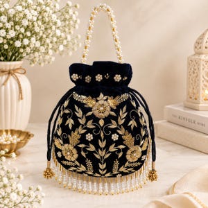 May include: A navy blue velvet drawstring pouch bag featuring intricate gold floral embroidery, sequin accents, and white pearl embellishments. The bag includes a pearl-beaded handle and a bottom fringe made of hanging white pearls and gold beads.