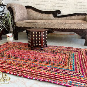 May include: A colourful, handwoven rug made from recycled materials. The rug has a rectangular shape and features a variety of colours, including red, pink, blue, green, and yellow. The rug is shown in a living room setting with a sofa and a small table.