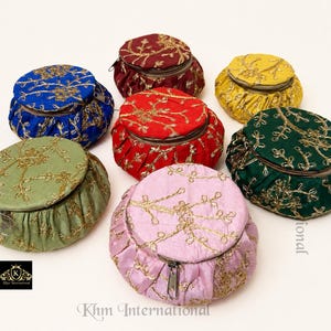 Op de afbeelding: Zeven ronde etuis met rits in blauw, bordeauxrood, rood, geel, groen, lichtgroen en roze. Elk etui is voorzien van goudkleurig borduurwerk in een rankenpatroon over de bovenkant en zijkanten. Een klein zwart logo met de tekst Khm International is zichtbaar.