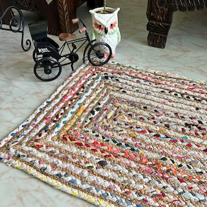 May include: A white, rectangular jute rug with a braided design. The rug is made with colorful fabric scraps woven into the jute. The rug is on a white marble floor.