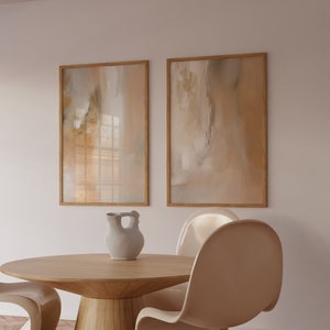 May include: Two framed abstract paintings in beige and brown tones hang on a white wall above a round wooden table with a white ceramic pitcher and four beige chairs.