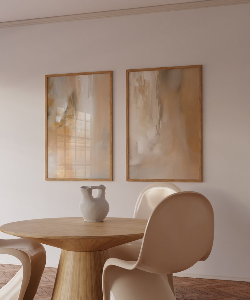 May include: Two framed abstract paintings in beige and brown tones hang on a white wall above a round wooden table with a white ceramic pitcher and four beige chairs.