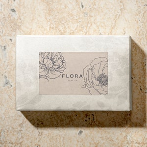 Puede incluir: Una caja blanca con una etiqueta marr&oacute;n que presenta un dibujo de l&iacute;nea de dos flores y el texto "FLORA SOAP CO."